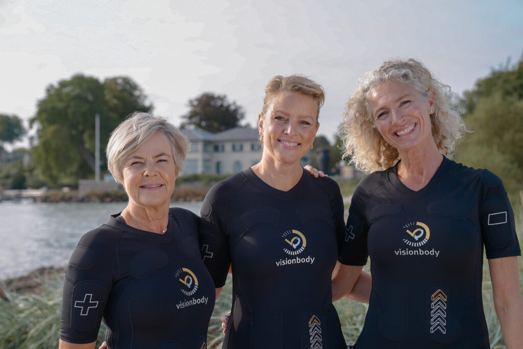 Tre glade kvinder i Visionbody EMS dragter smiler til kameraet ved vandet.